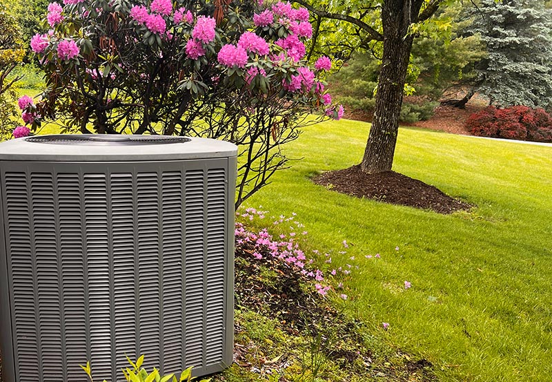 A grey outdoor central air conditioning condenser unit positioned in a beautifully landscaped yard.