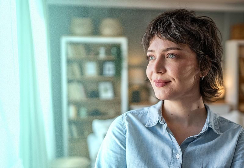 A close-up of a woman with short, wavy dark hair and light blue eyes, wearing a light-colored chambray button-up shirt.